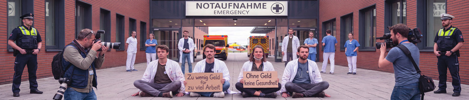 Ärztinnen und Ärzte der „Allerletzten Generation“ kleben sich vor Notauf nahme der neuen Klink CURE4ALL fest. Sie fordern mehr Geld und kürzere Ausbildung.